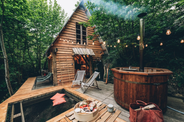 Vu de prêt terrasse de cabane avec transates, une table, jacuzzi, lit de toile et cabane en bois en forêt.